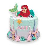 Disney Princess Ariel Cake featuring the Little Mermaid Princess Ariel amid fondant shells, colourful vibrant mermaid tails made of white chocolate, and edible pearls on a turquoise-pink buttercream frosting base.